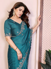 Woman wearing a teal saree with silver embroidery against a light pink wall.