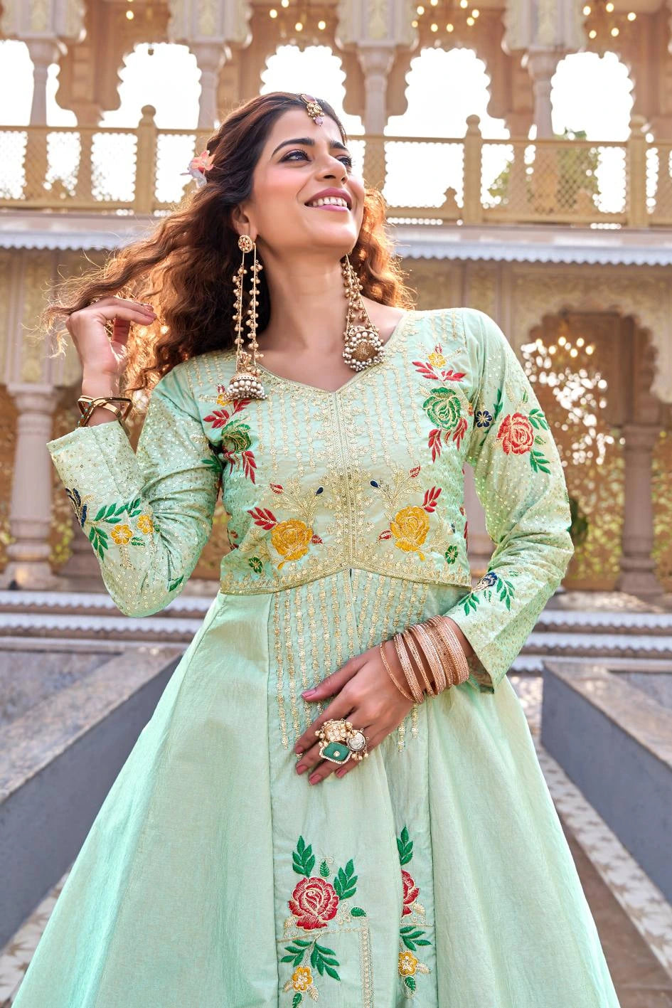 Woman in a light green traditional outfit with floral embroidery standing in front of an architectural background.