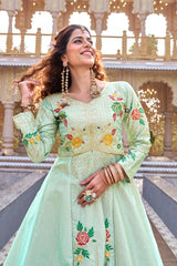 Woman in a light green traditional outfit with floral embroidery standing in front of an architectural background.