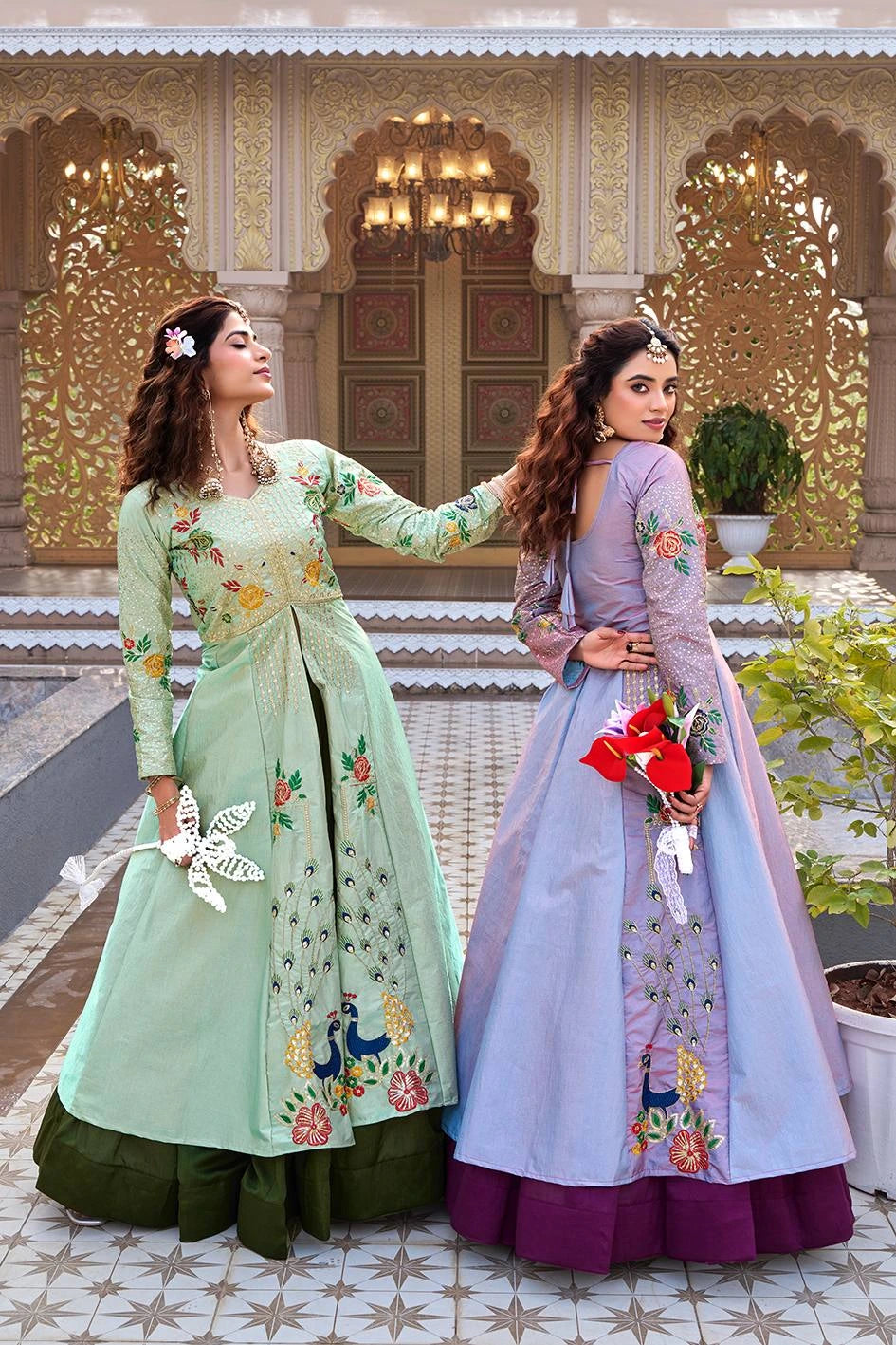 Two women in traditional outfits standing in front of an ornate architectural background.