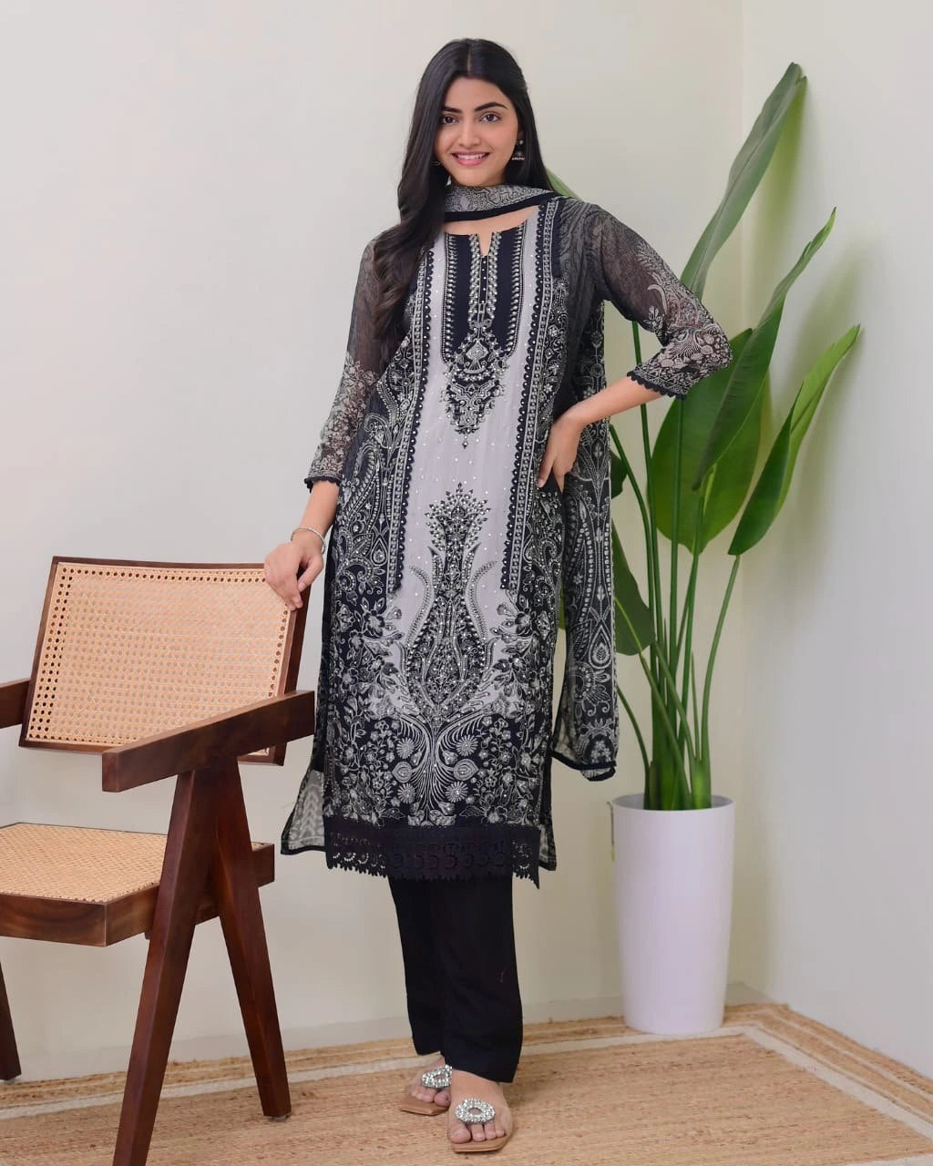 Woman wearing a black and white embroidered outfit in a room with a chair and plant.