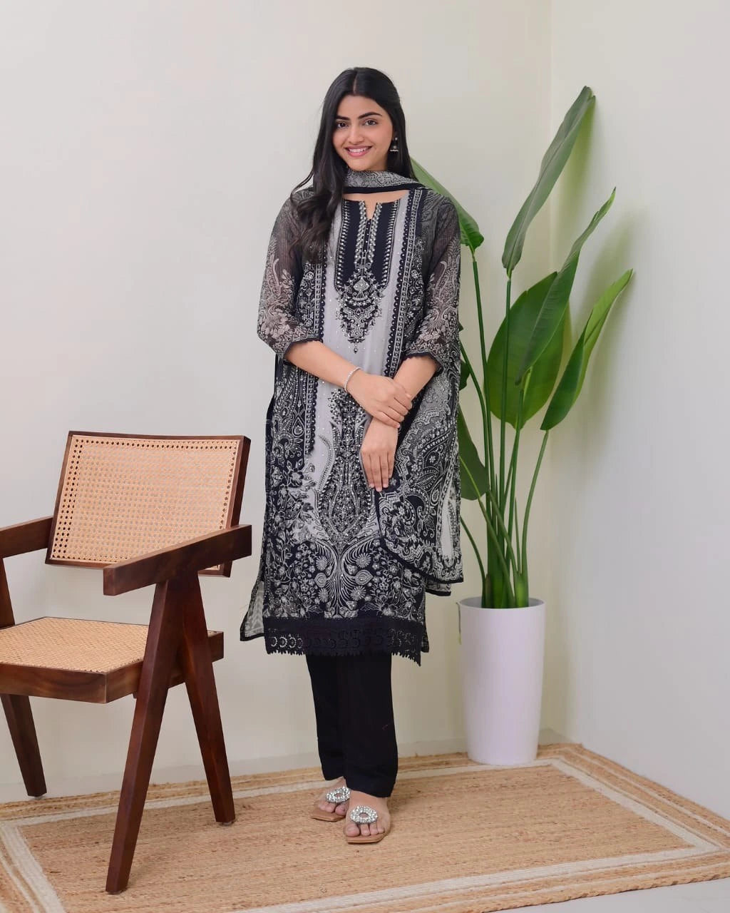 Woman in a black and white patterned outfit standing in a room with a chair and plant.