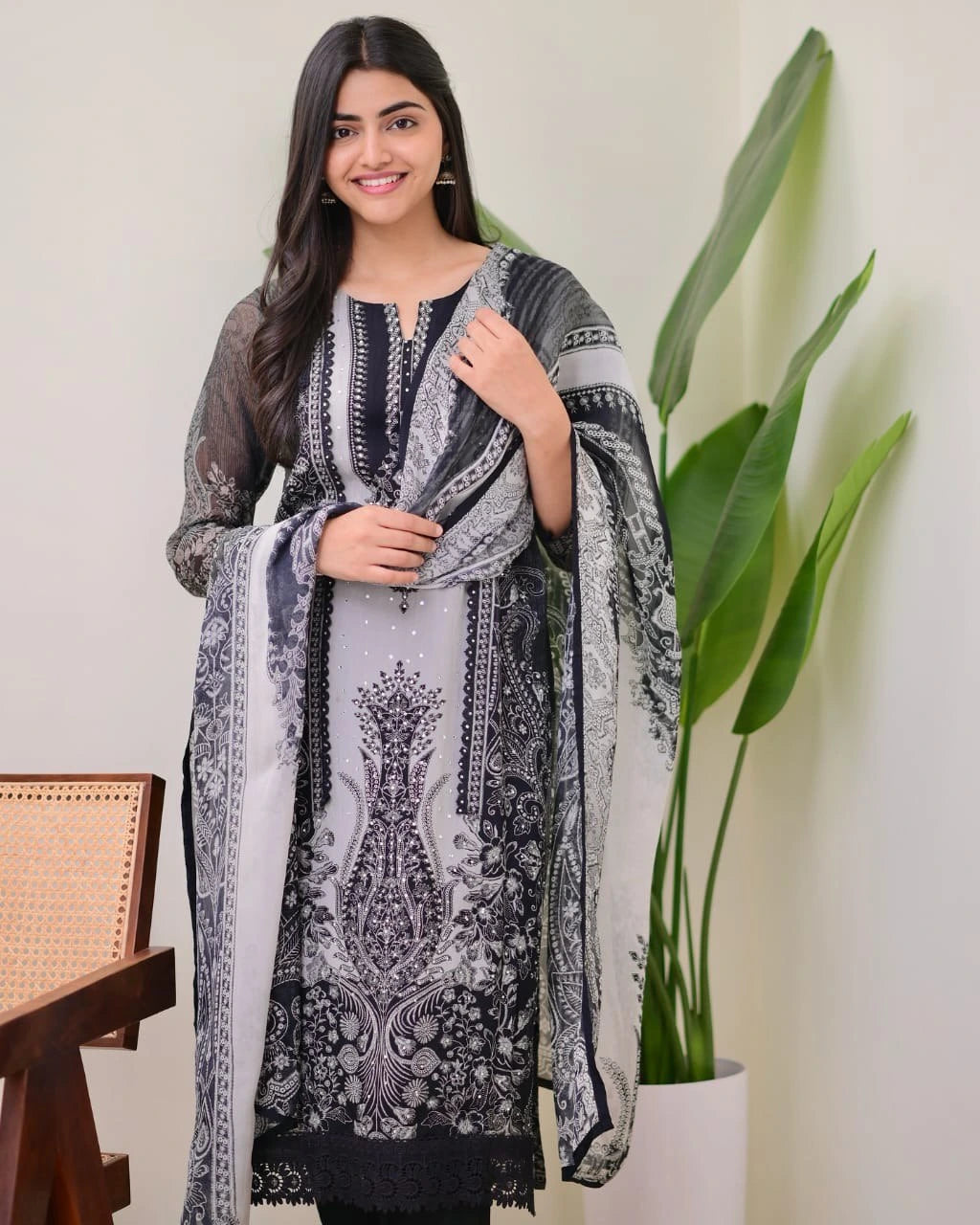 Woman wearing a black and white patterned dress with a scarf, standing indoors with a plant in the background.