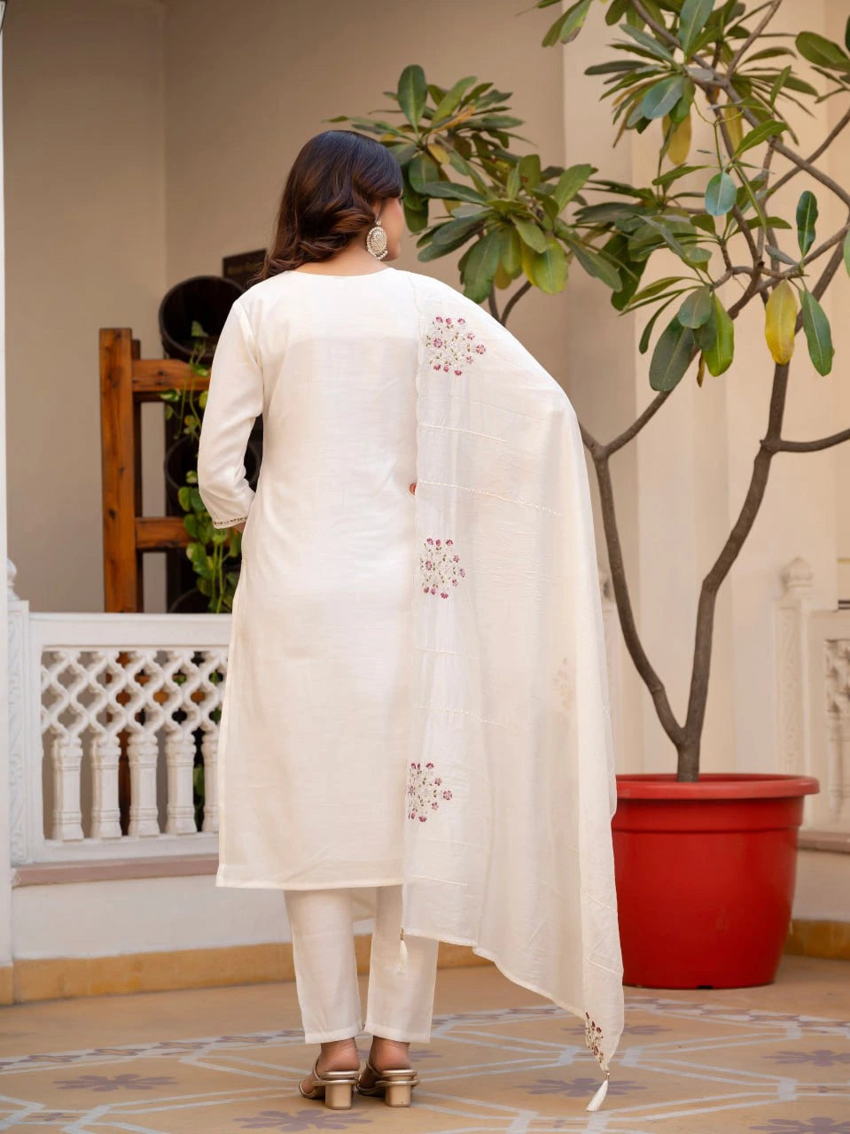 Woman wearing a white embroidered outfit standing in a room with a plant and decorative elements.