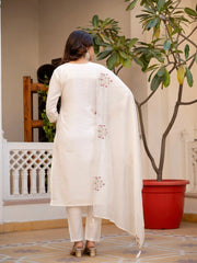 Woman wearing a white embroidered outfit standing in a room with a plant and decorative elements.