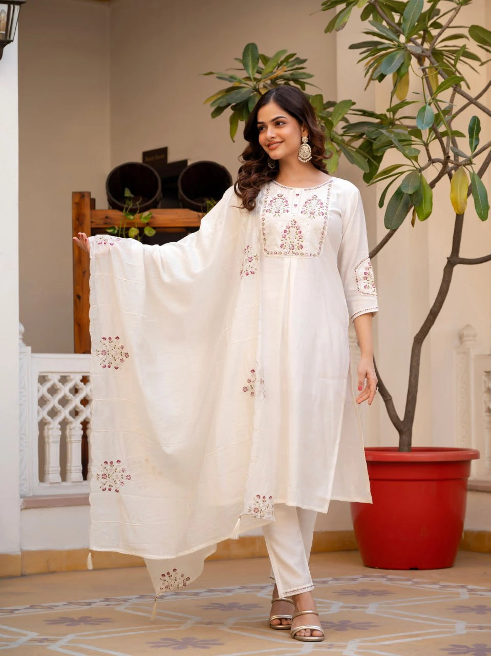 Woman in a white embroidered outfit standing indoors with plants in the background