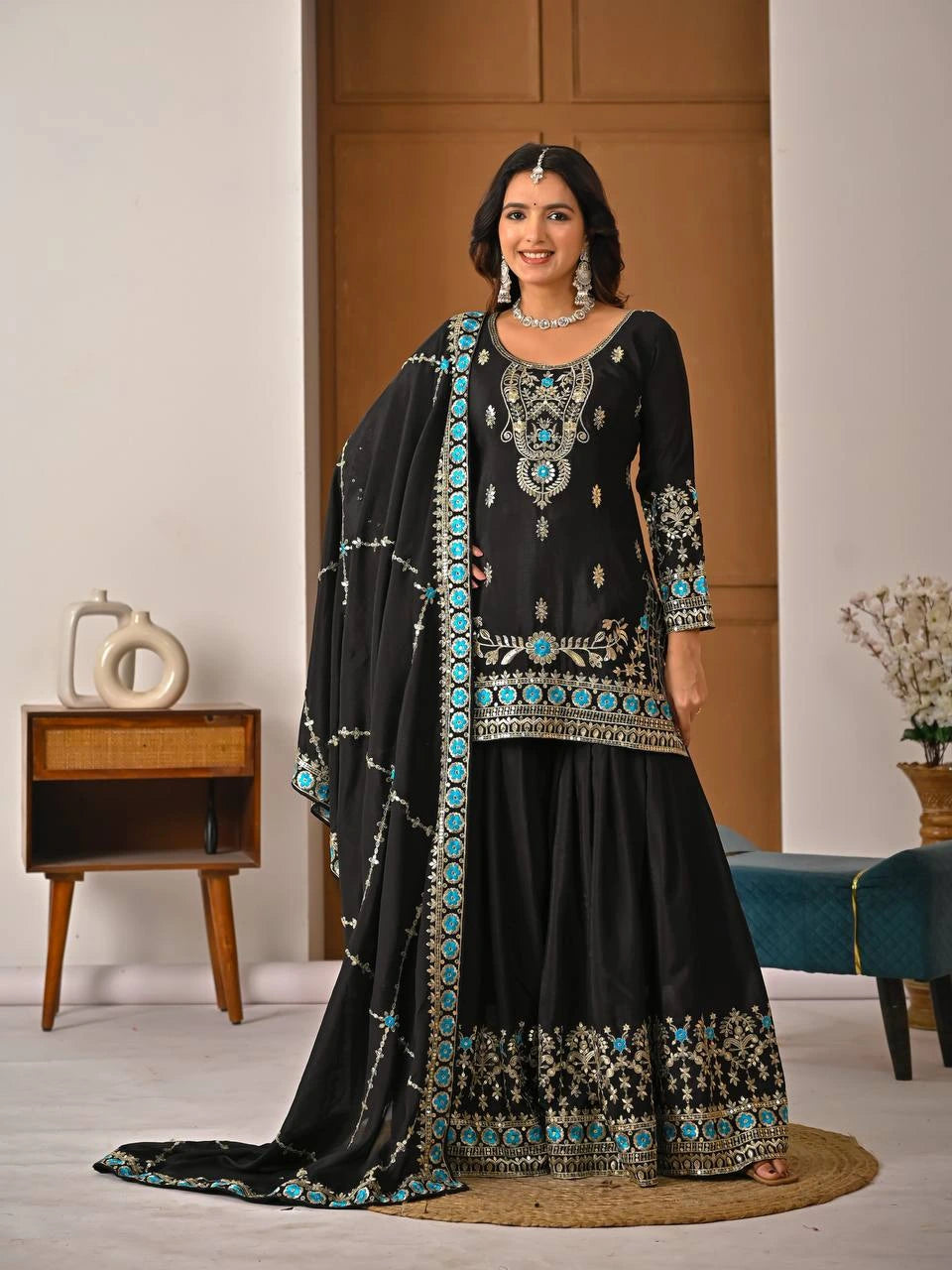 Woman wearing a black embroidered traditional outfit in a room with a wooden cabinet and decorative items.