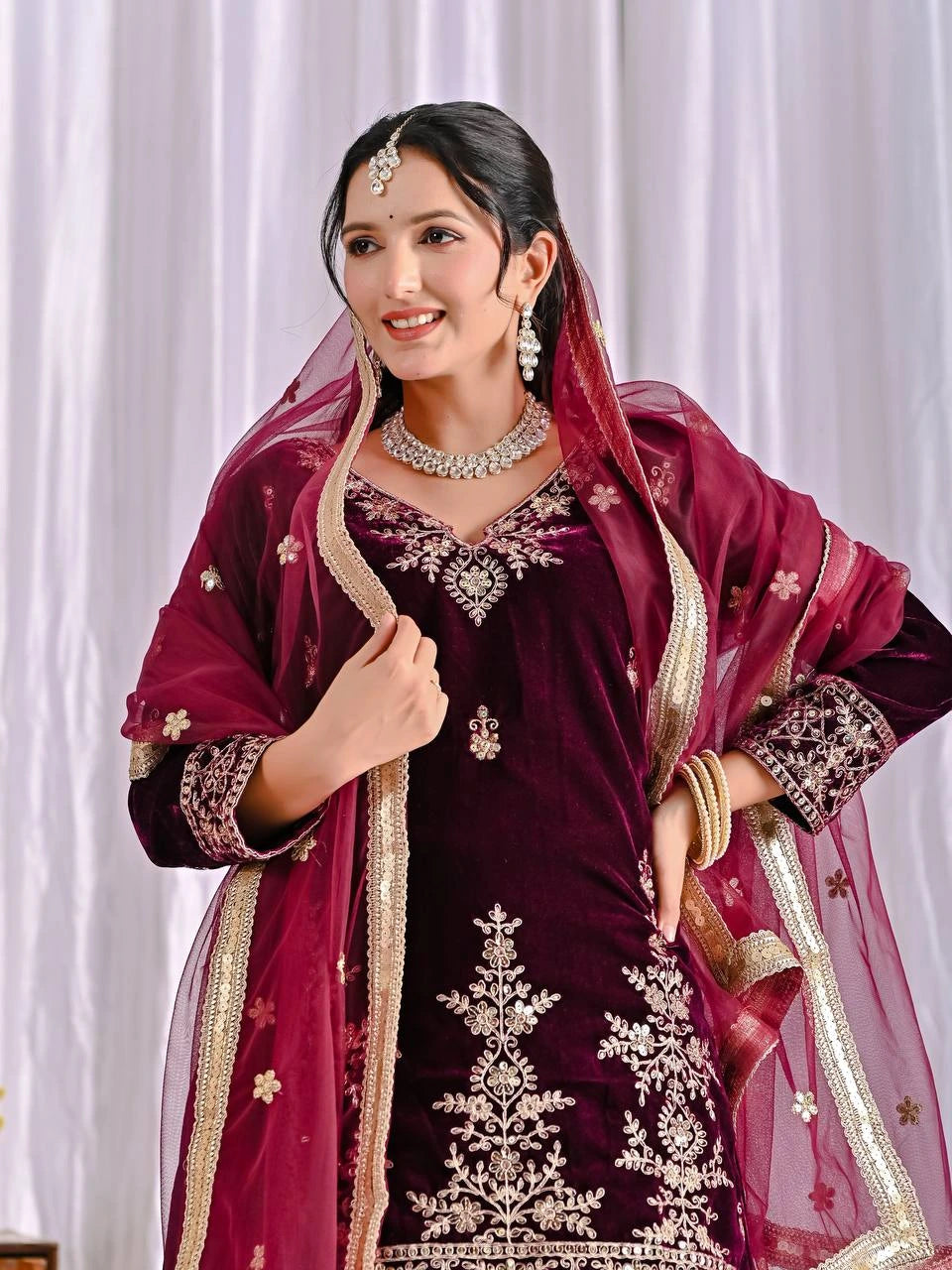 Woman in traditional maroon and gold embroidered outfit with a sheer dupatta against a white curtain background