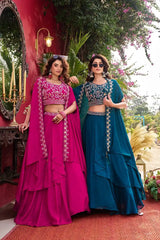 Two women in traditional pink and blue lehengas standing outdoors with a decorative background.