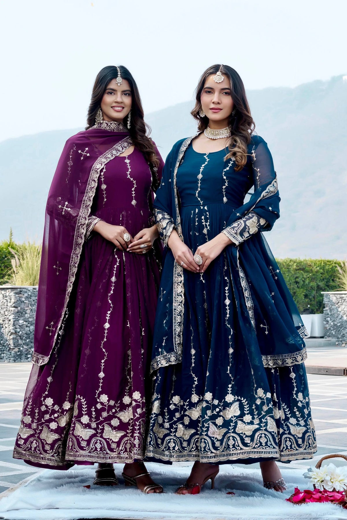 Two women in traditional embroidered dresses standing outdoors with mountains in the background.