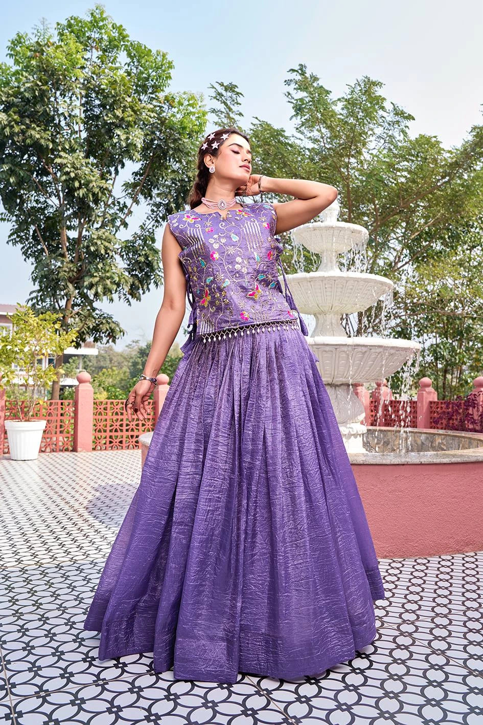 Woman in a purple traditional outfit standing outdoors with trees and a fountain in the background