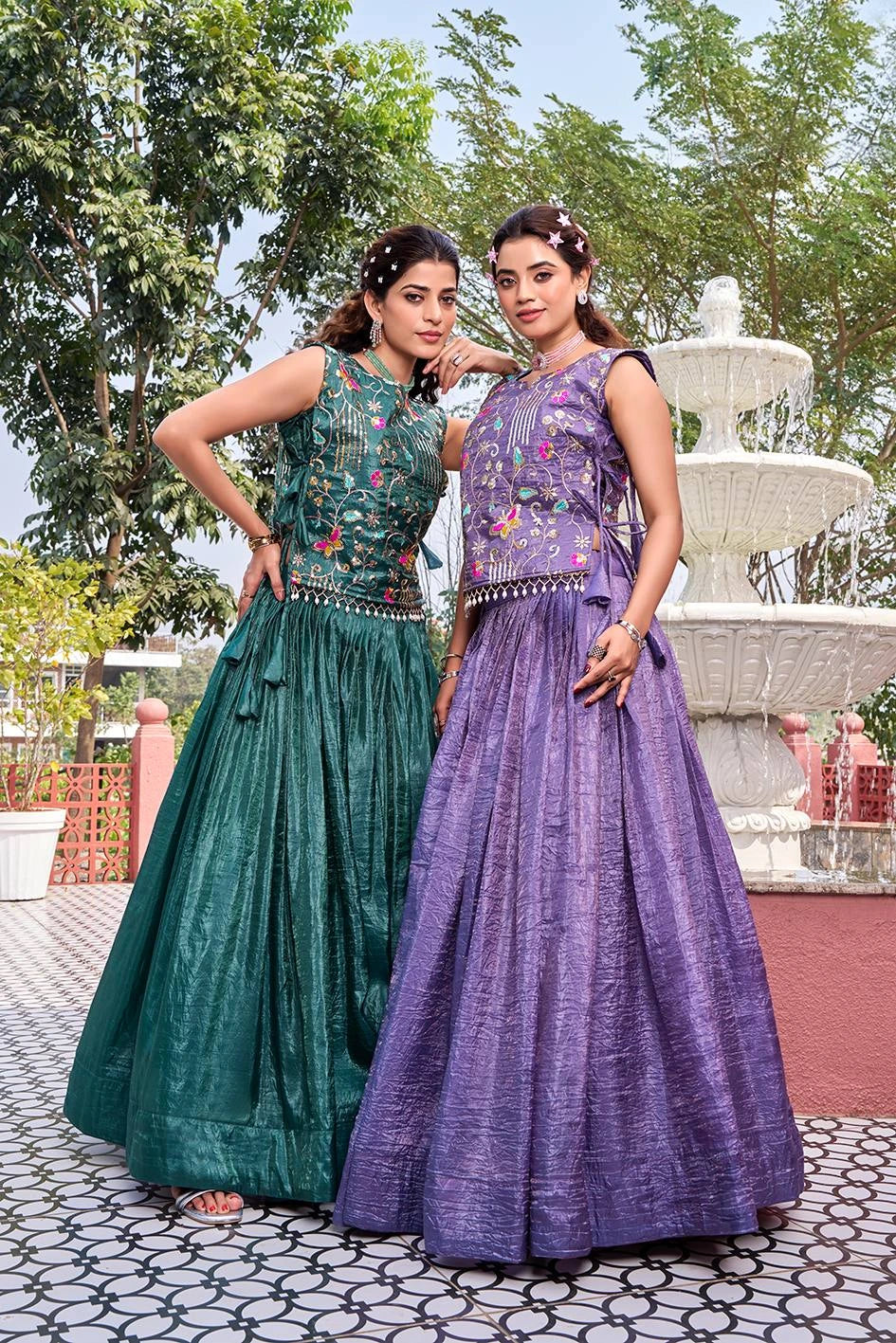 Two women in traditional dresses standing outdoors with a fountain in the background.