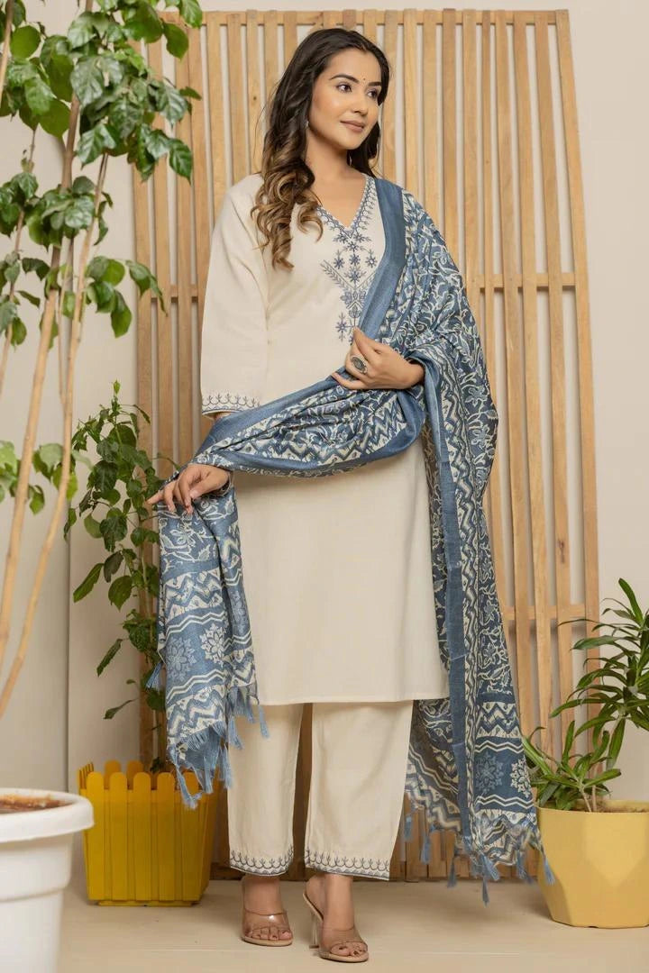 Woman in a cream outfit with a blue patterned scarf standing indoors with plants around.