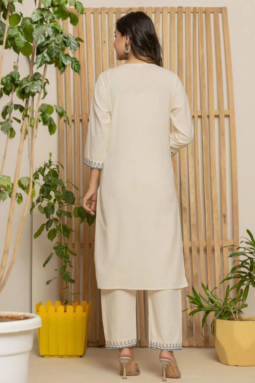 Woman in a beige kurta with white pants standing in a room with plants and a wooden screen.
