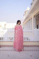 Woman in a pink saree standing on a balcony with a white building and blue sky in the background