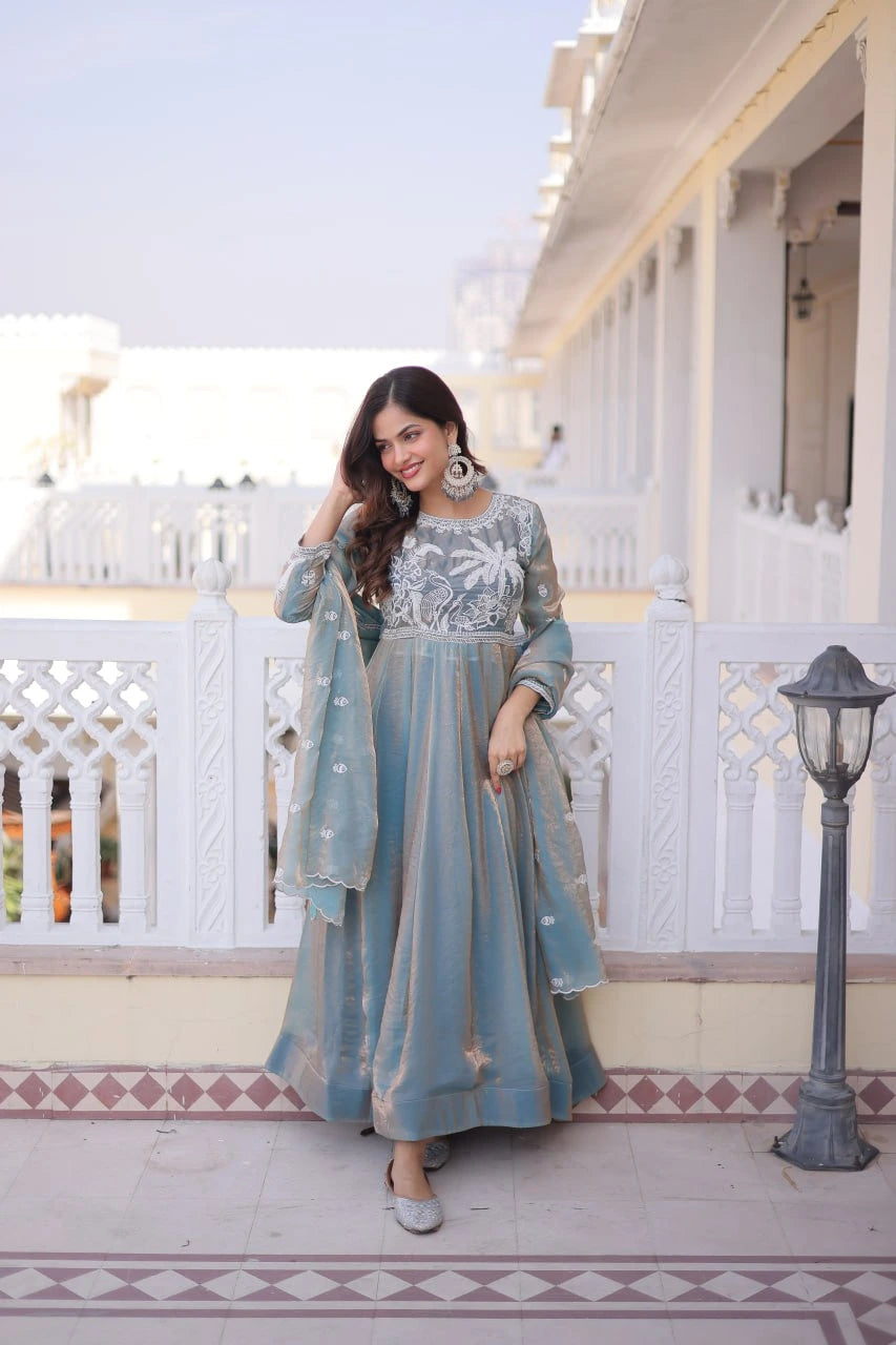 Woman in a light blue traditional outfit standing on a balcony with white railings and a building in the background.