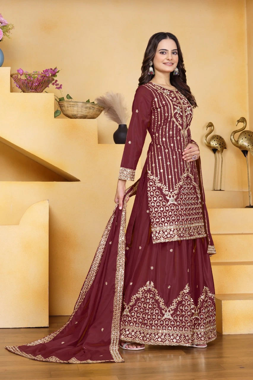 Woman in a maroon embroidered traditional outfit standing in a decorated room.