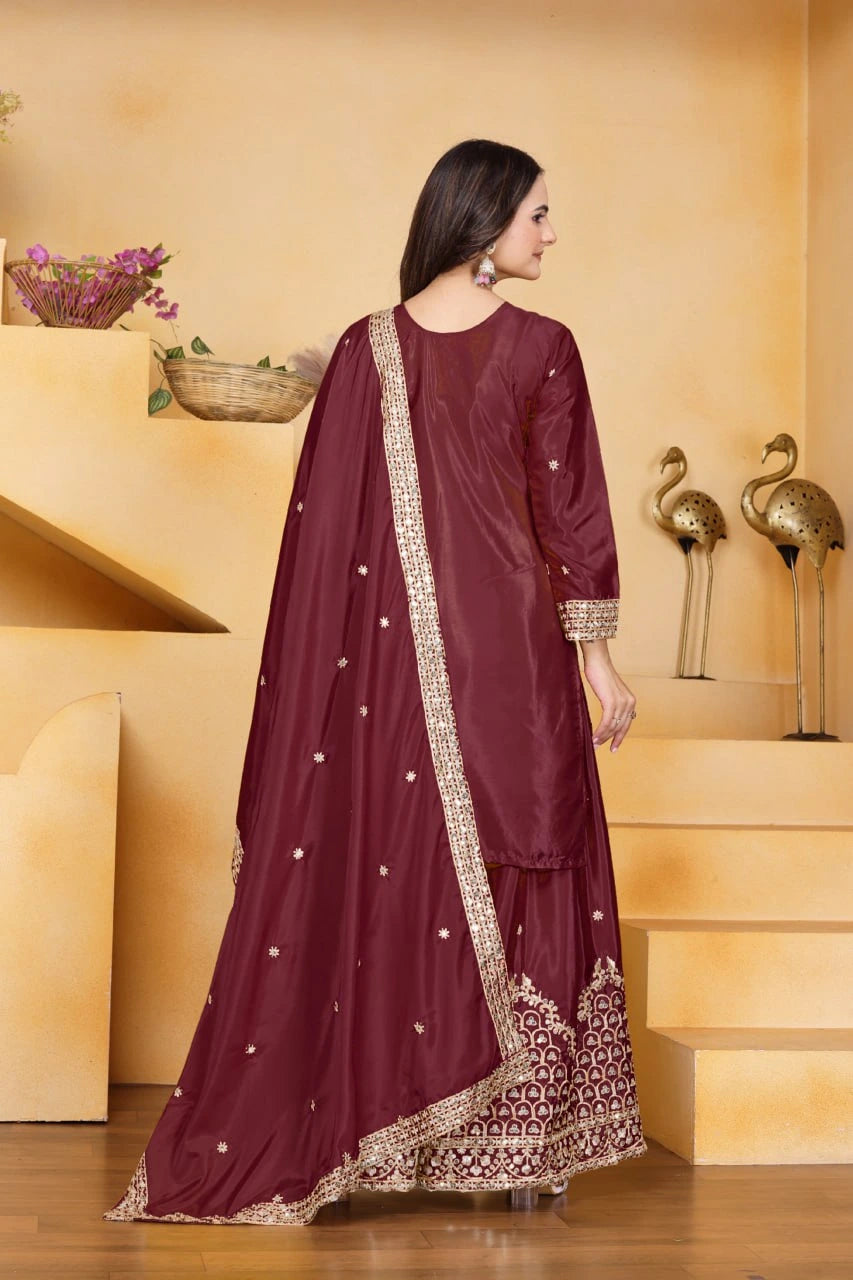 Woman in a maroon traditional outfit with gold embroidery standing in a decorative setting.