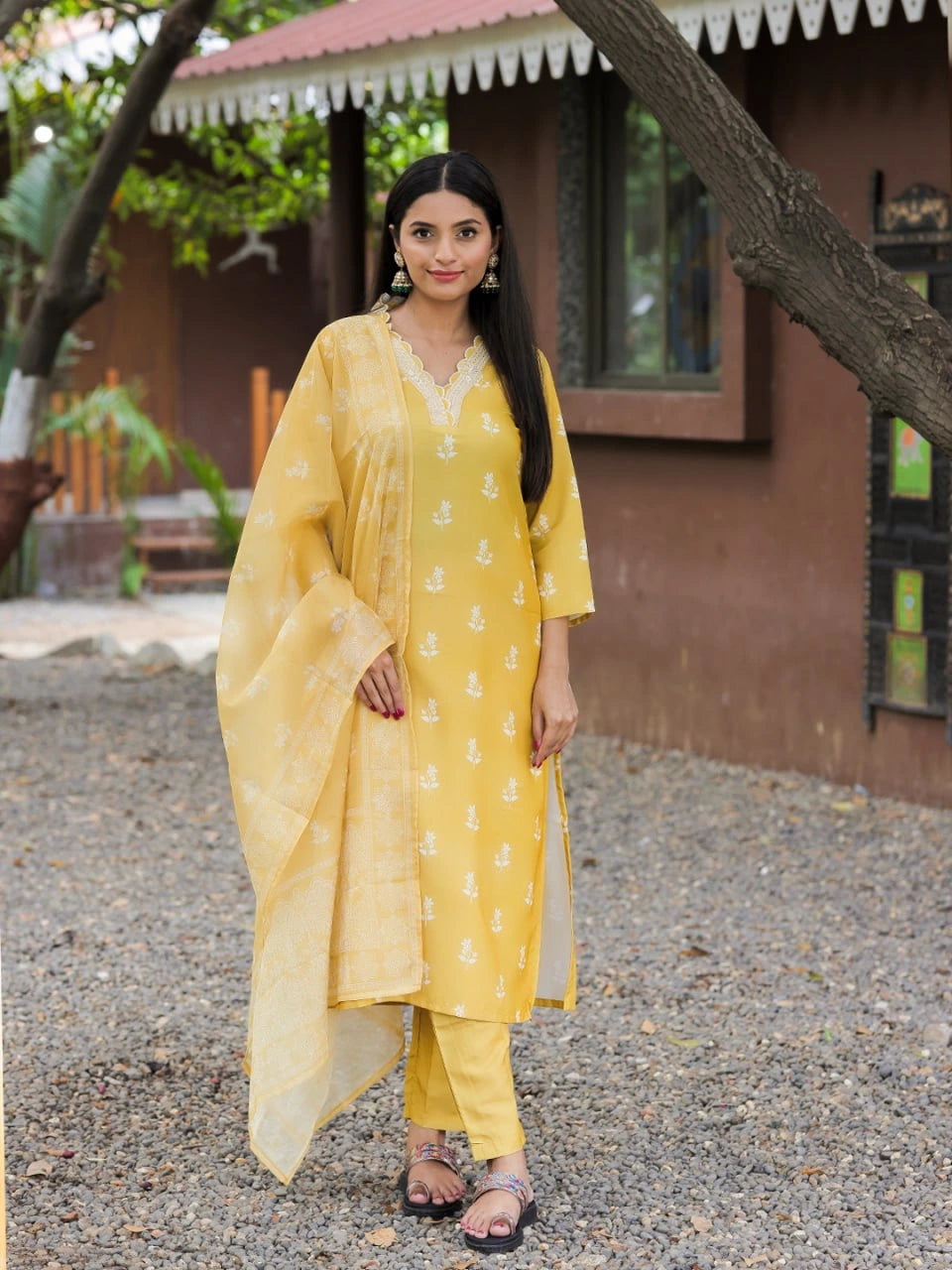 Woman in a yellow traditional outfit standing outdoors with a house and trees in the background