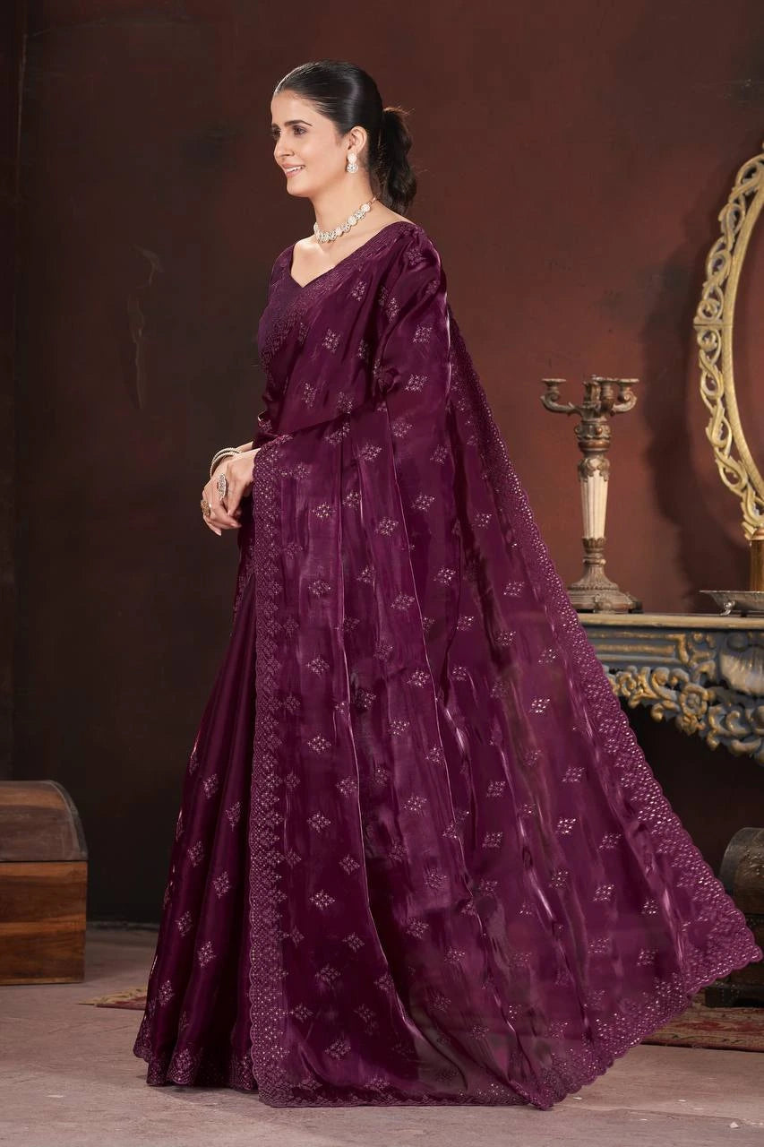 Woman in a long, dark purple embroidered dress standing against a brown background.