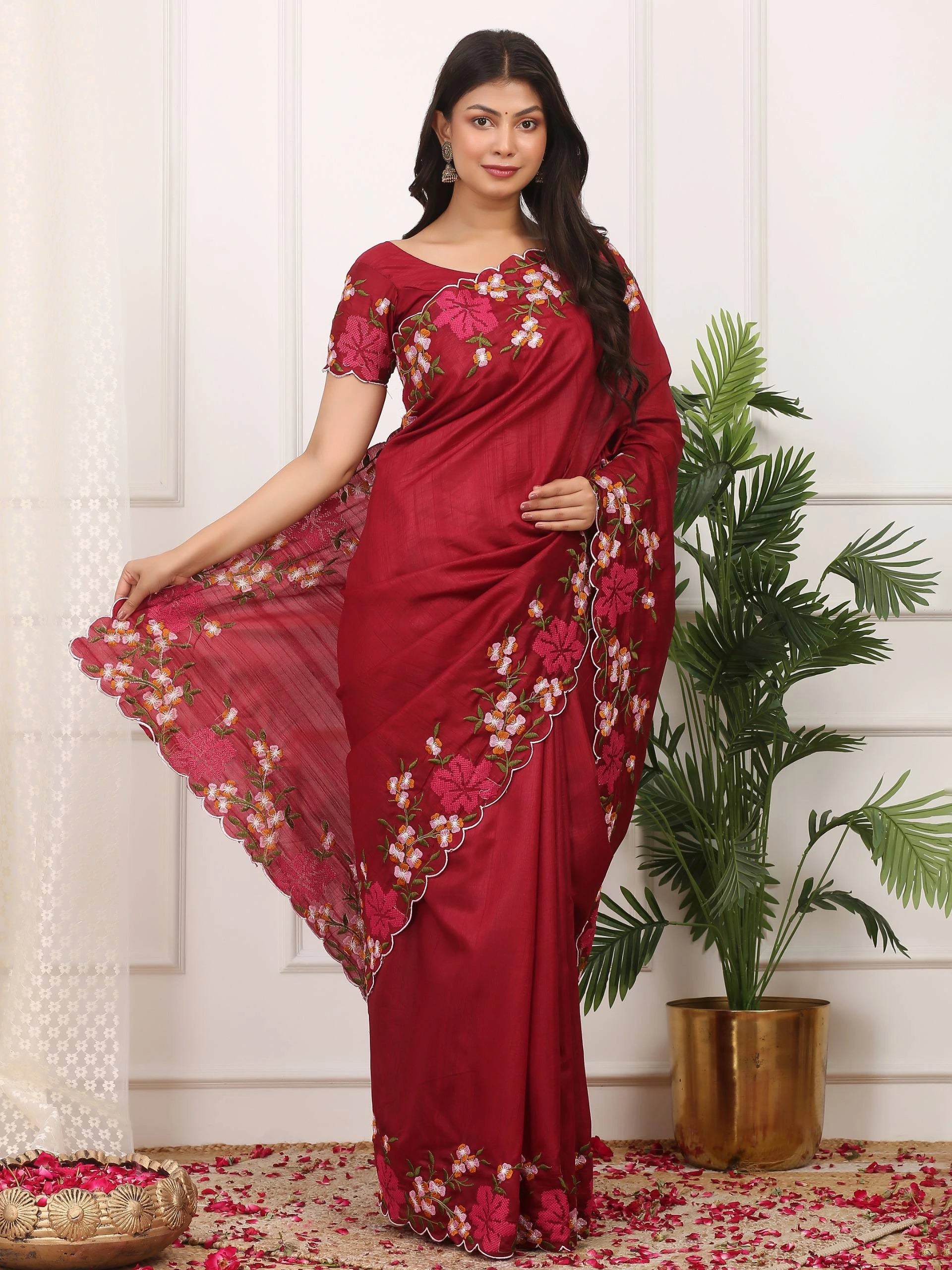 Woman wearing a red saree with floral patterns in a room with a plant and decorative items.