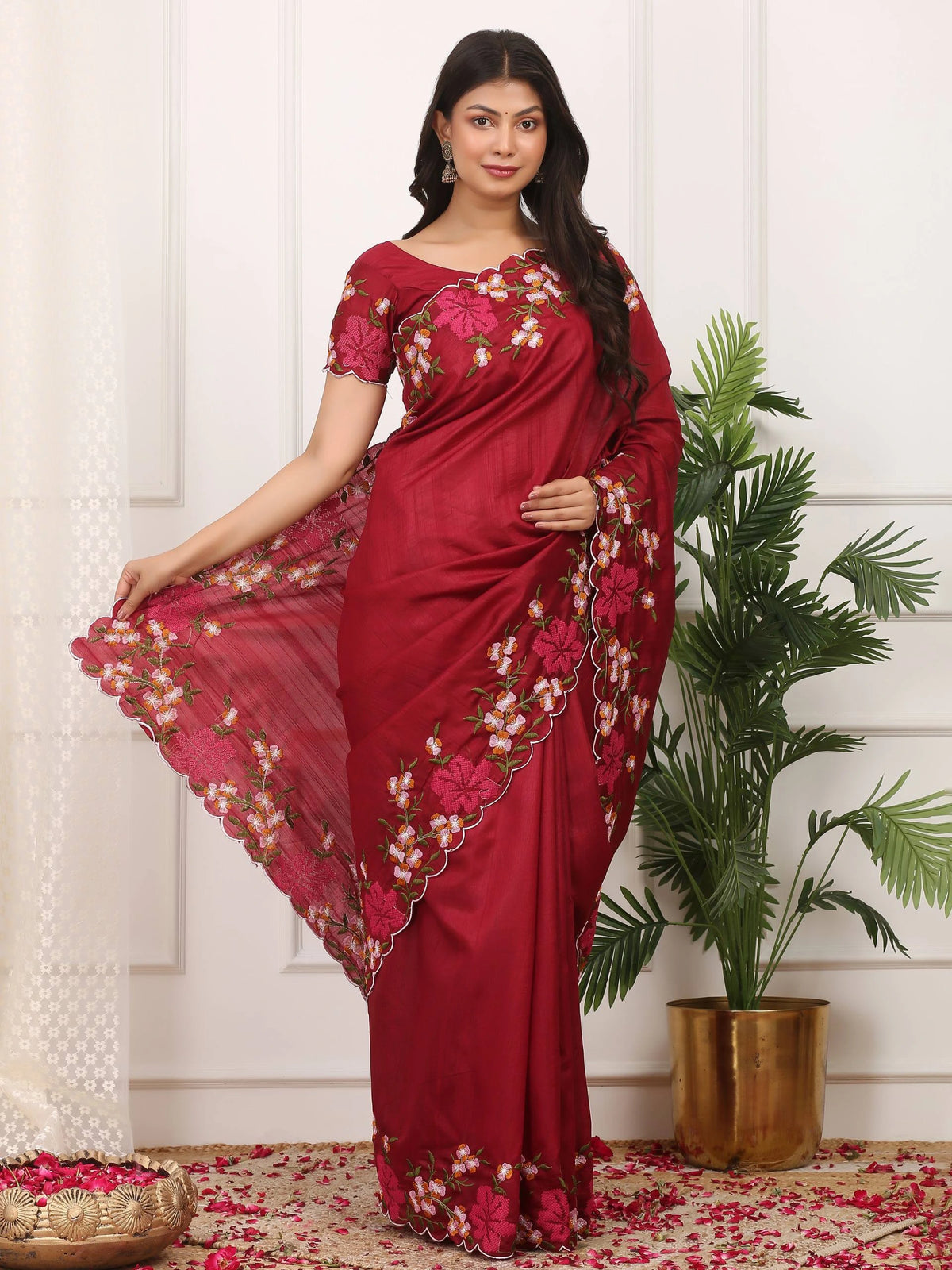 Woman wearing a red saree with floral patterns in a room with a plant and decorative items.