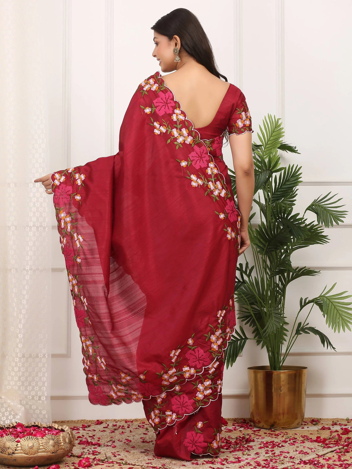 Woman wearing a red saree with floral patterns in a room with a plant and decorative items.