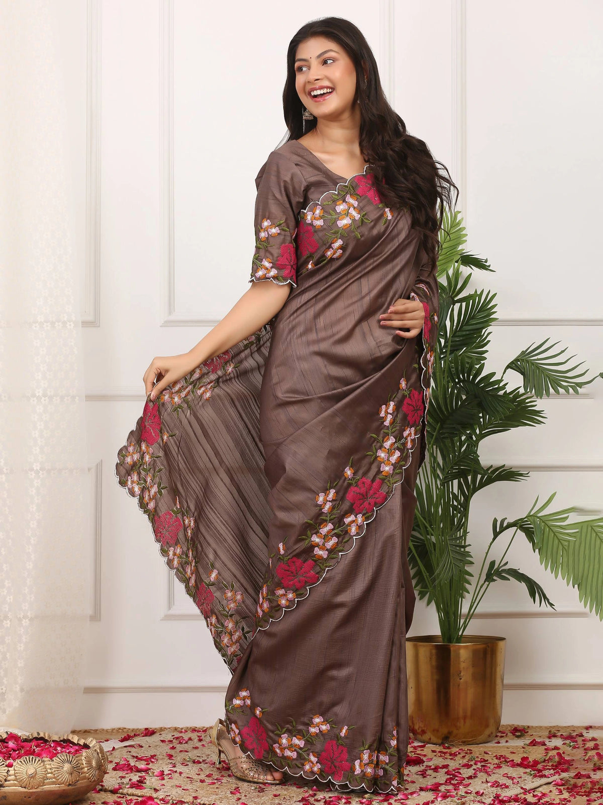 Woman wearing a brown saree with floral patterns in a room with a plant and decorative items.