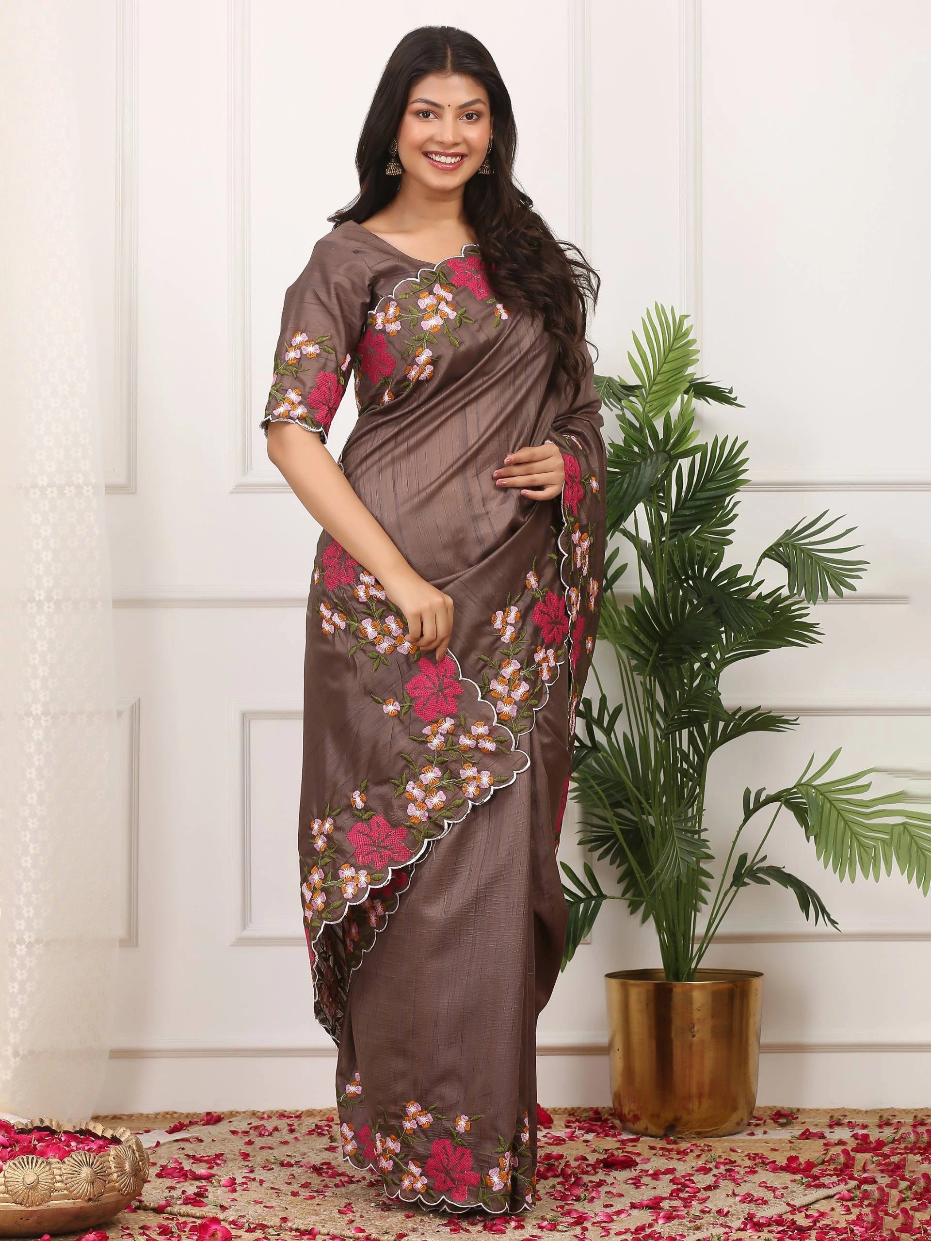 Woman wearing a brown saree with floral patterns standing next to a plant indoors.