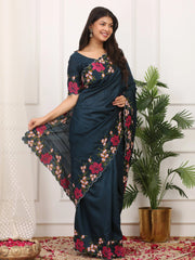 Woman wearing a dark blue saree with floral patterns in a room with a plant and decorative items.