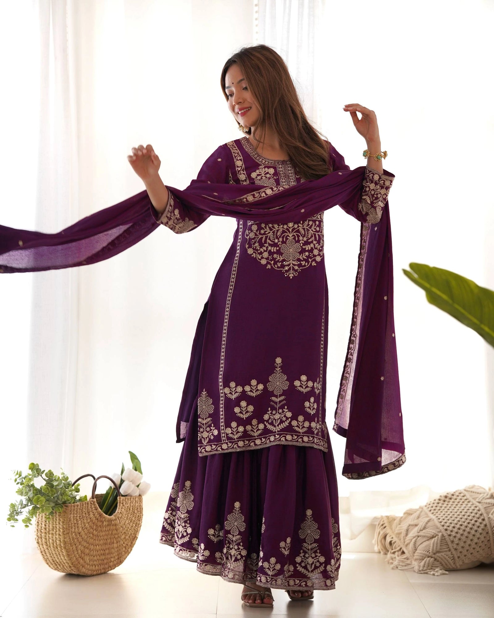 Woman wearing a purple embroidered traditional outfit with a matching dupatta, standing indoors.