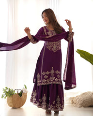Woman wearing a purple embroidered traditional outfit with a matching dupatta, standing indoors.