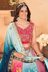 Woman in traditional pink and blue embroidered outfit with jewelry, sitting indoors.
