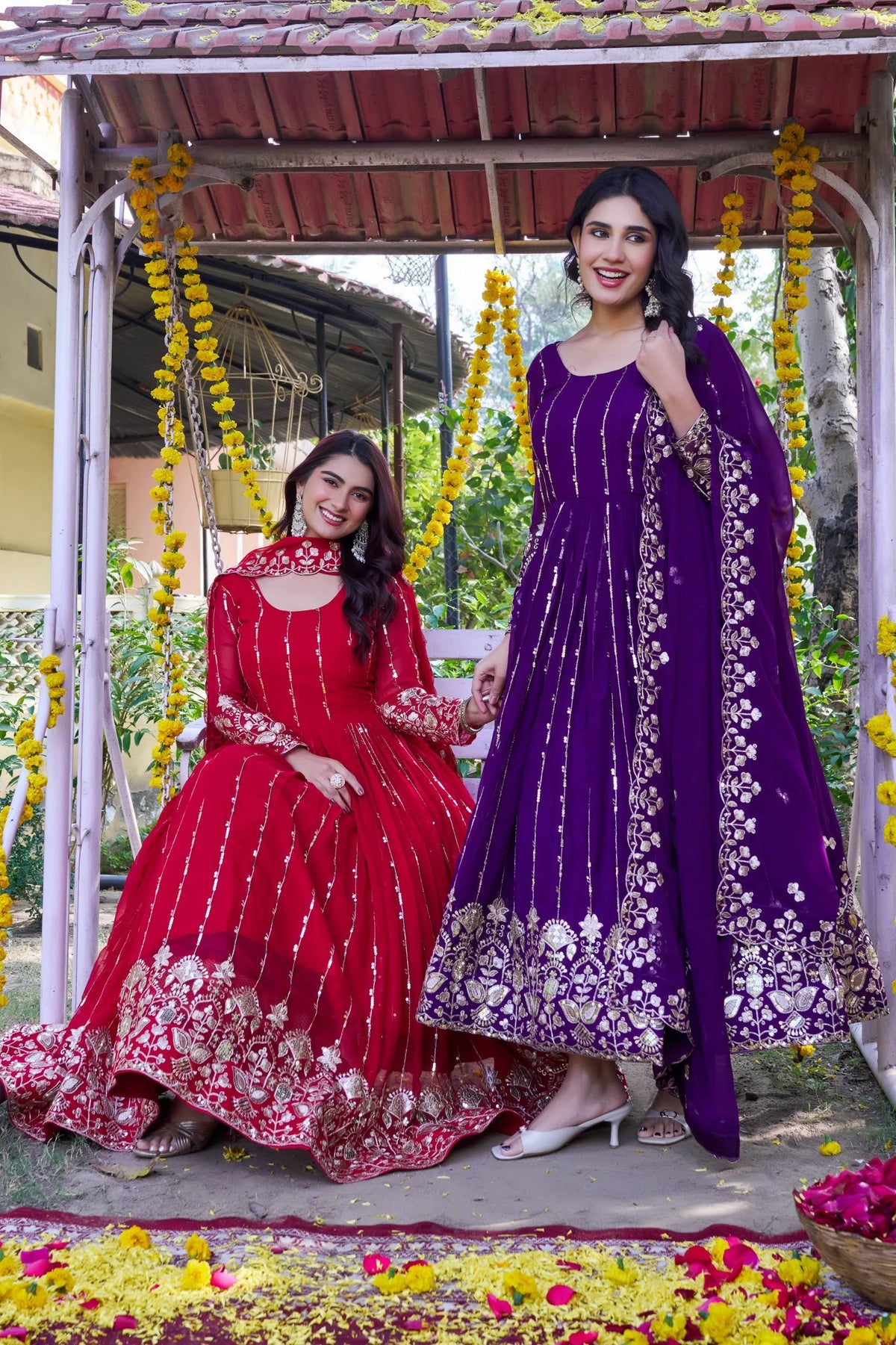 Two women in traditional red and purple dresses sitting on a swing decorated with flowers.