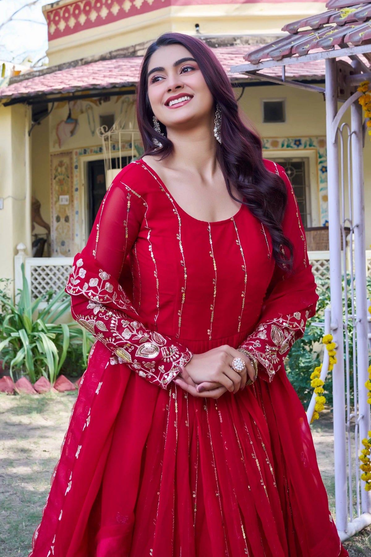 Woman in a red traditional outfit standing outdoors with a building and garden in the background
