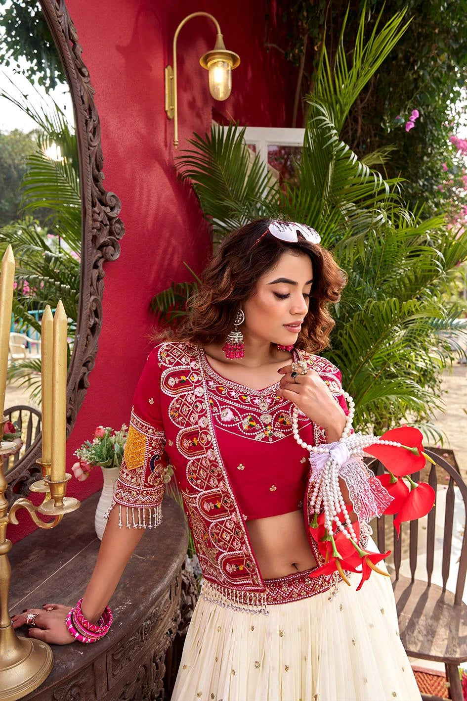 Woman in traditional red and white outfit standing in a decorative outdoor setting with plants and candles.