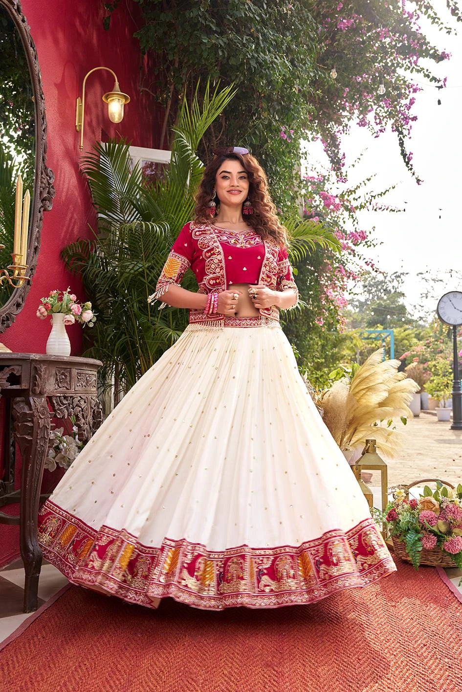 Woman in a traditional outfit with red and white colors standing in an outdoor setting with plants and flowers.