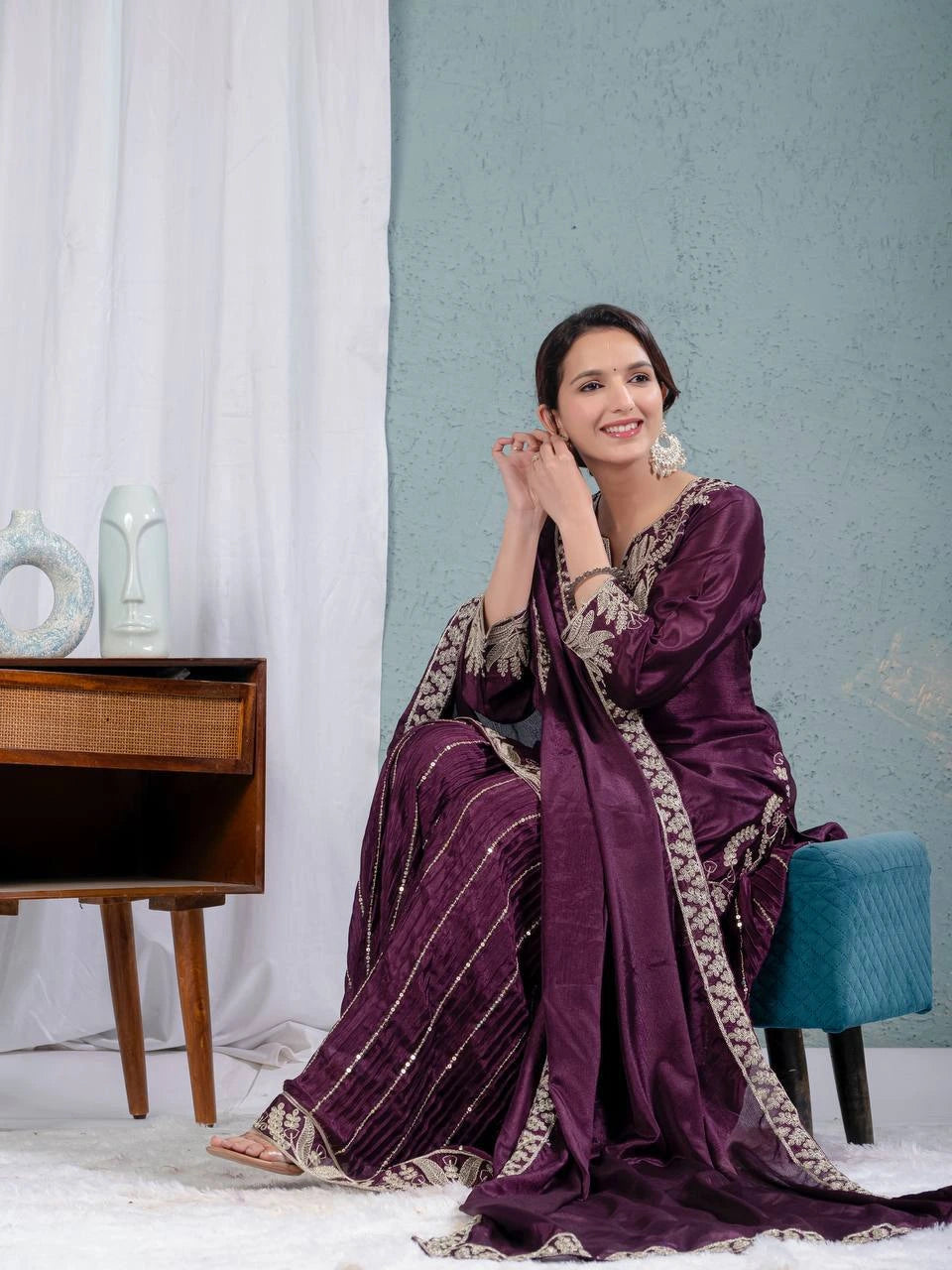 Woman in a purple traditional outfit sitting in a room with a blue wall and wooden furniture.