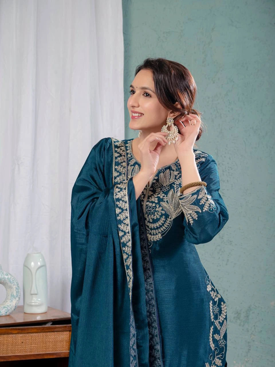 Woman wearing a blue embroidered dress adjusting her earrings indoors.