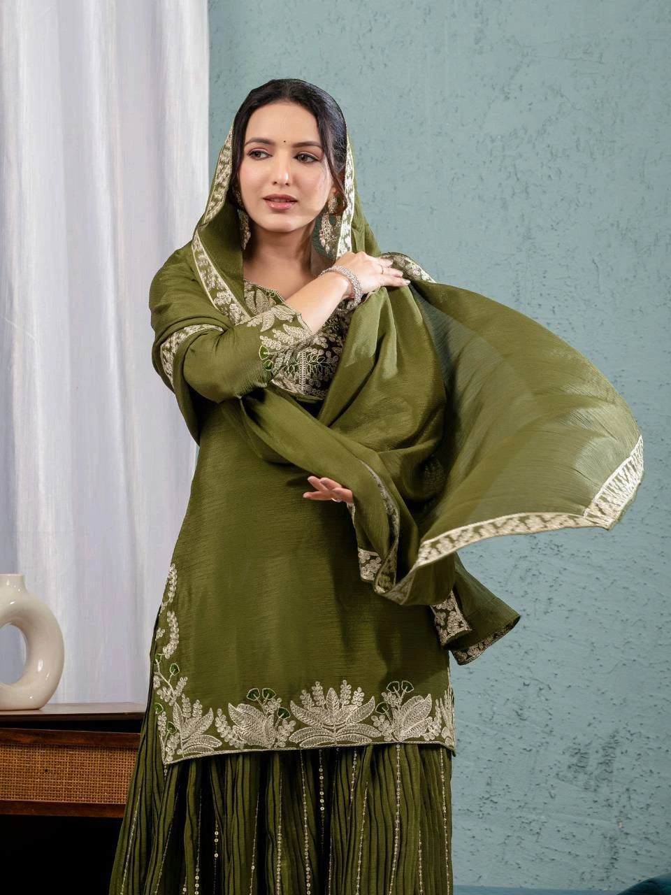 Woman wearing a green traditional outfit with a matching dupatta against a light blue wall.