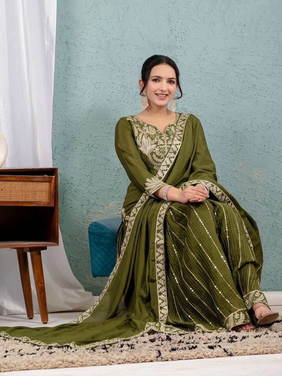 Woman in a green traditional outfit sitting on a rug with a light blue wall and wooden furniture in the background.