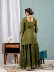 Woman in a green traditional outfit standing in a room with a blue wall and wooden side table.