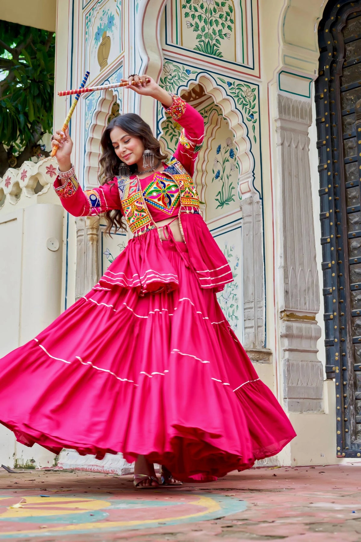 Woman in a bright pink traditional outfit with colorful patterns, standing in front of a decorative archway.