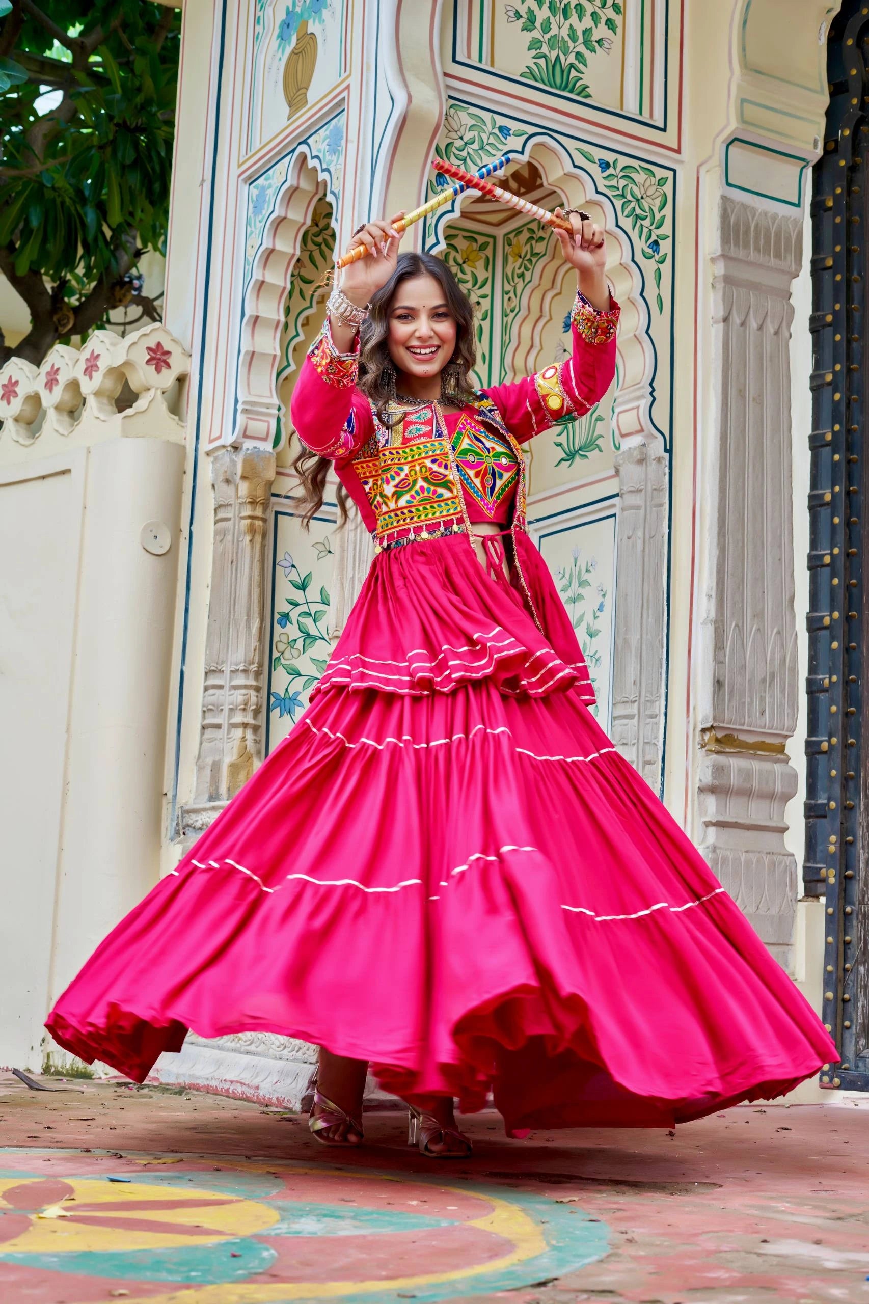 Woman in a bright pink traditional outfit with colorful patterns, dancing in front of an ornate wall.
