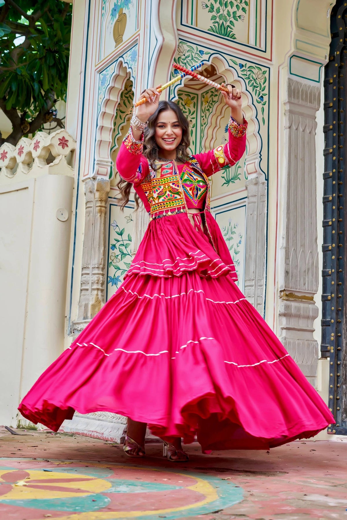 Woman in a bright pink traditional outfit with colorful patterns, dancing in front of an ornate wall.