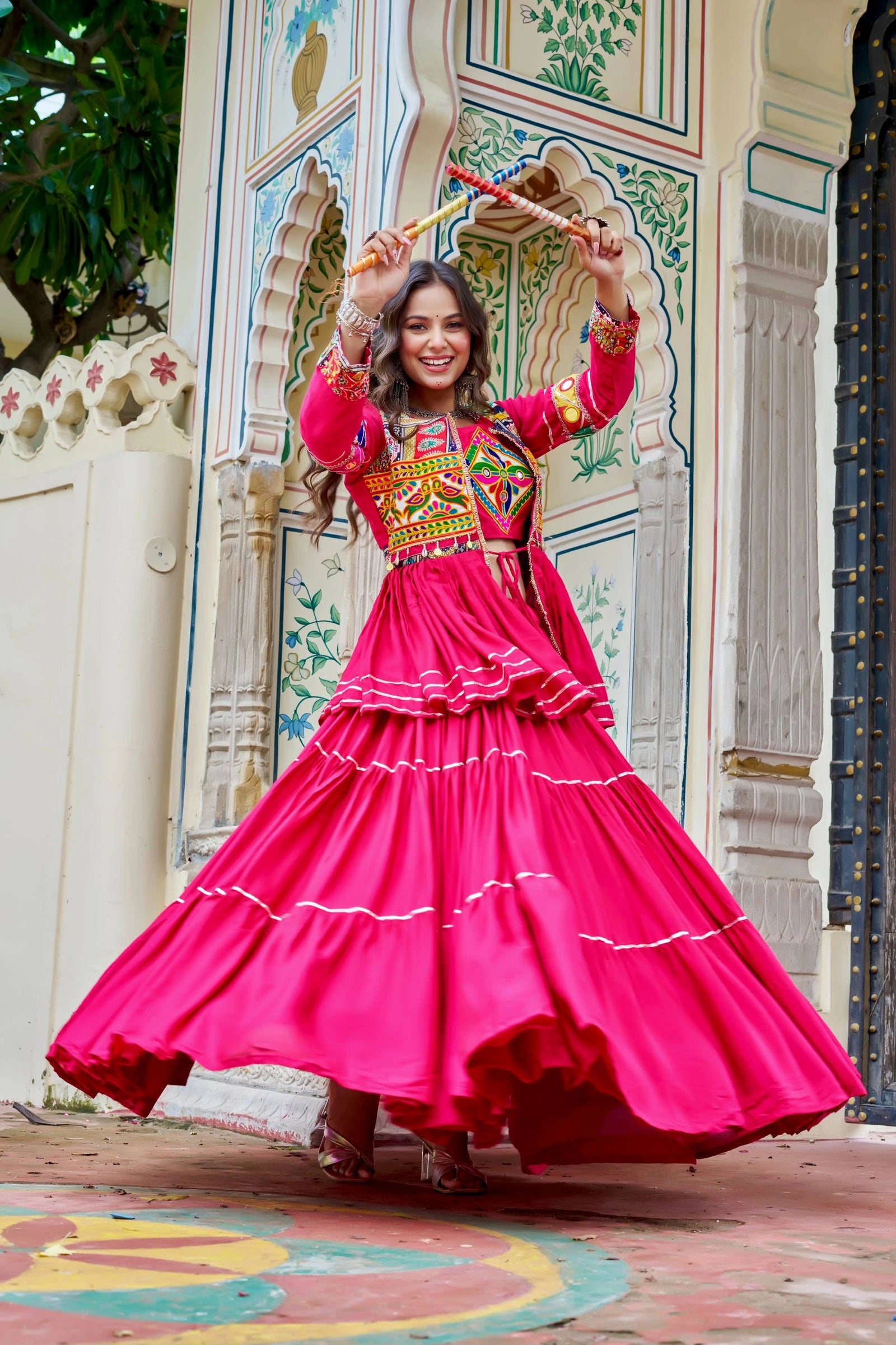 Woman in a bright pink traditional outfit with colorful patterns, dancing in front of an ornate wall.
