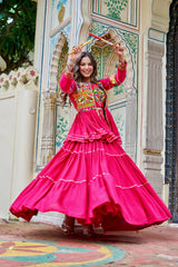 Woman in a bright pink traditional outfit with colorful patterns, dancing in front of an ornate wall.