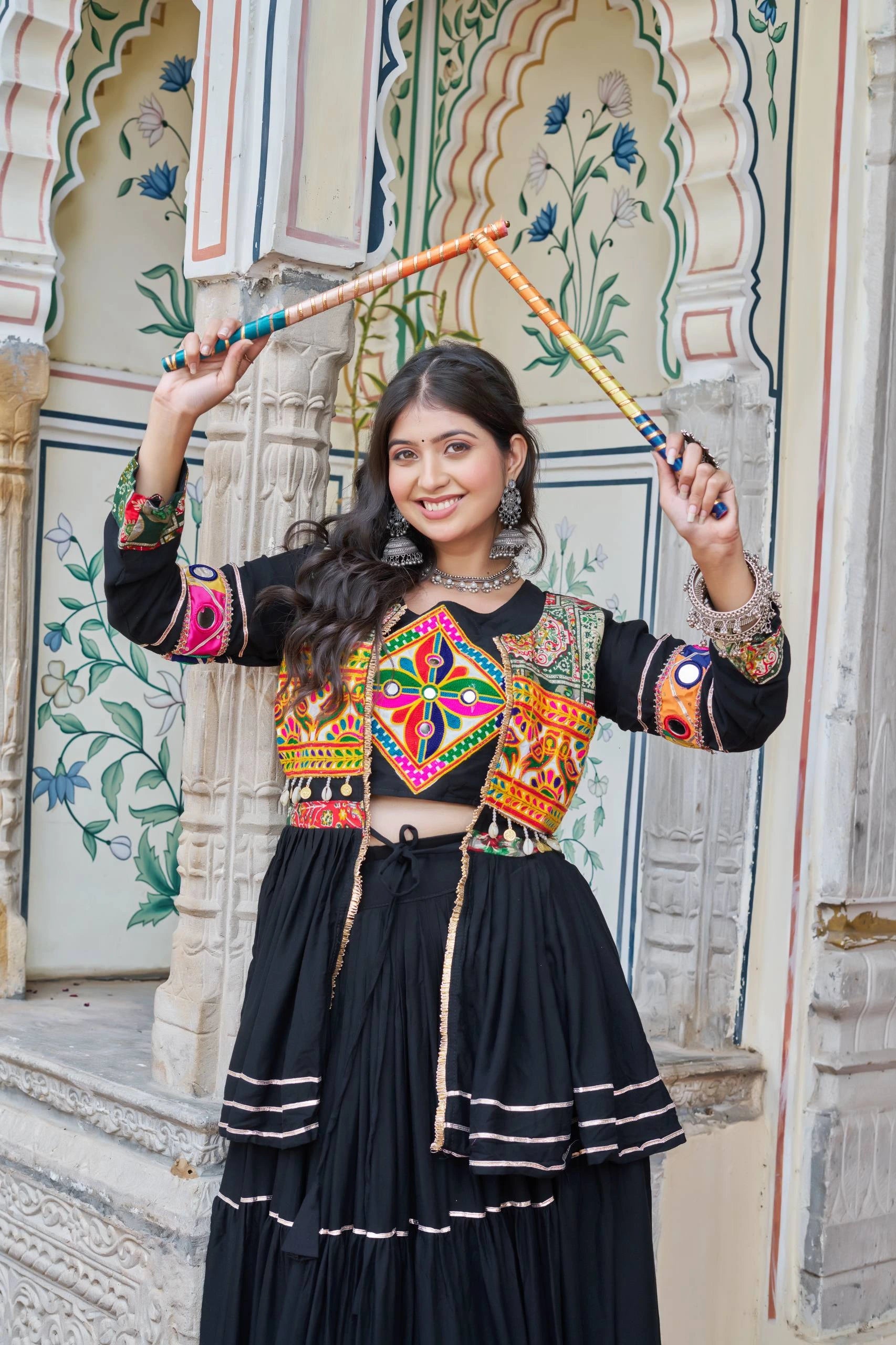 Woman in traditional outfit with colorful embroidery holding a decorative staff, standing in front of ornate architectural background.
