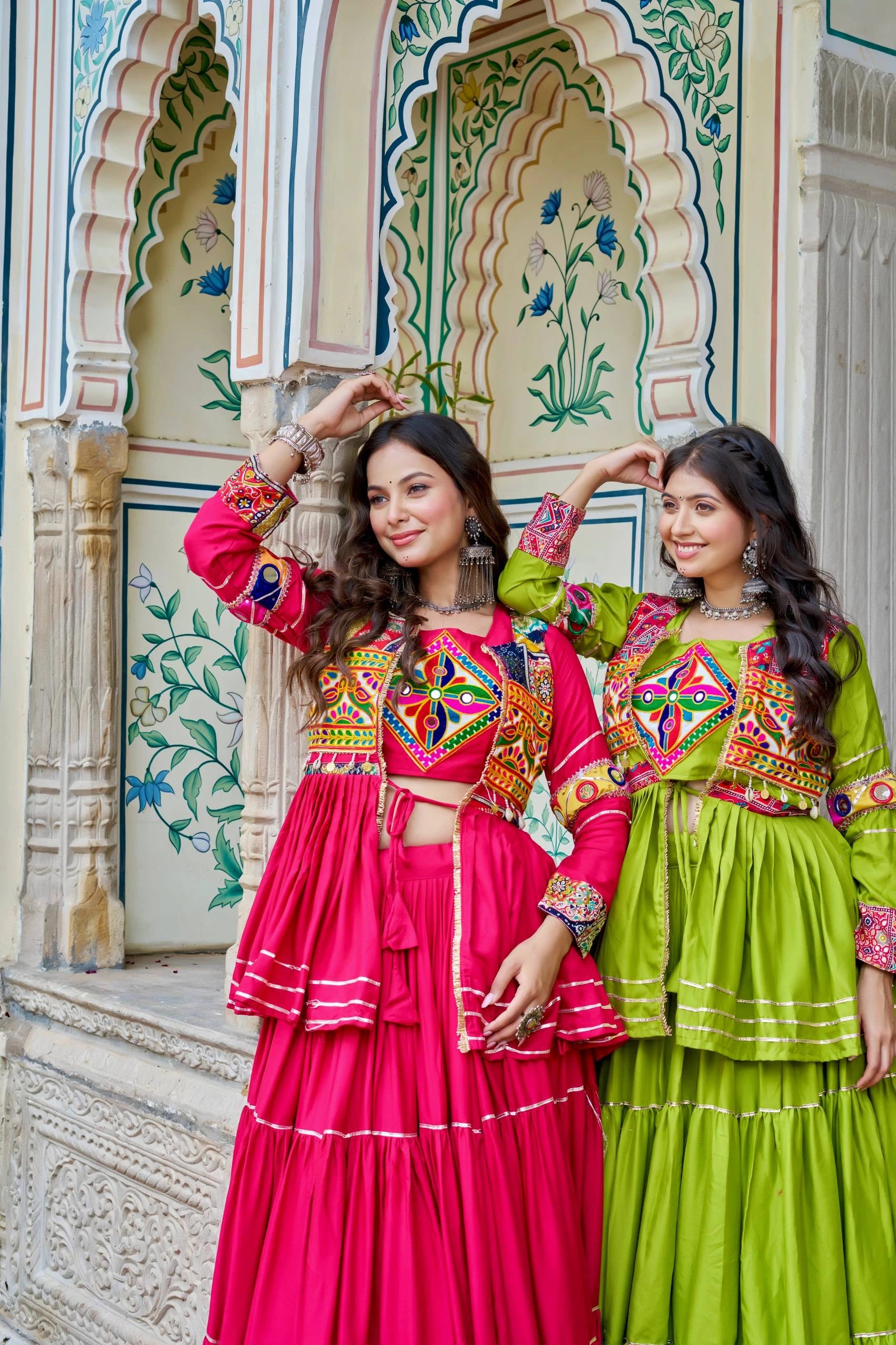 Two women in colorful traditional outfits posing in front of an ornate architectural background.
