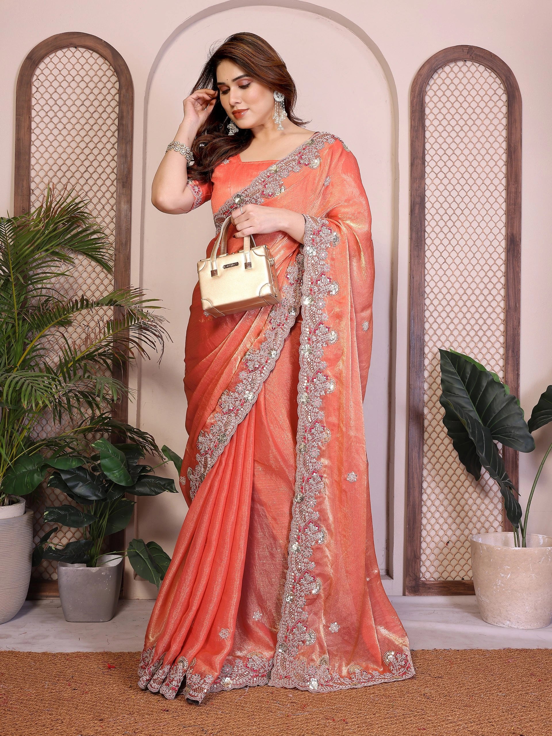 Woman in an orange saree with silver border holding a gold handbag indoors.